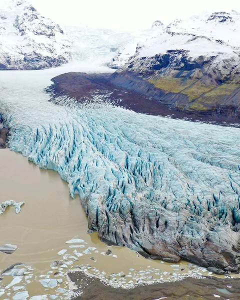 Lengua de glaciar agrietada vista desde el aire, entre montañas nevadas, con témpanos en la laguna de su frente