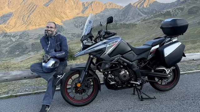 Luis sentado en una valla de madera junto a una Suzuki V-Strom, con las montañas iluminadas por el sol de la tarde