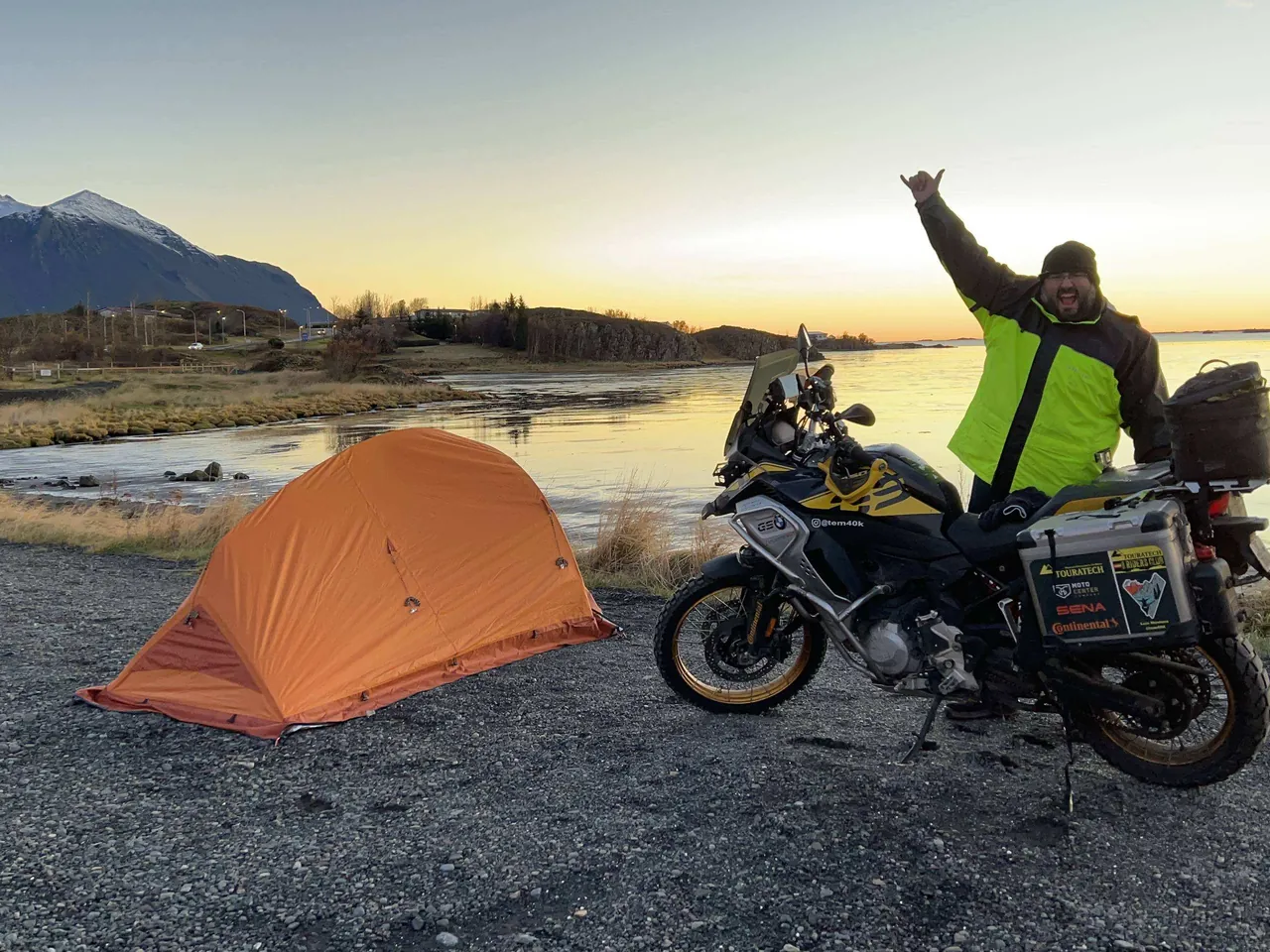 Luis levanta el brazo junto a su BMW F850 GS Adventure y la tienda naranja, al atardecer en Islandia